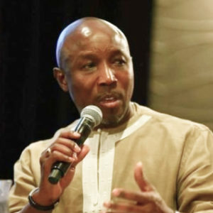 A photo of Magnus Conteh, holding a microphone while speaking, wearing a beige jacket